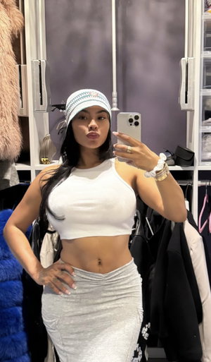 Woman in white crop top and gray skirt poses in closet, wearing beanie, holding phone.
