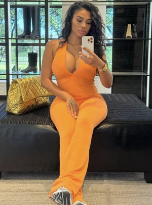 Woman in orange jumpsuit sits on black ottoman, holding phone for mirror selfie indoors.