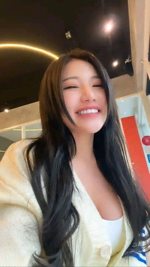 Asian woman with long dark hair in yellow cardigan taking selfie indoors.