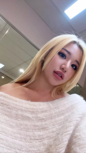 Blonde woman in off-shoulder sweater, indoor setting, close-up selfie.