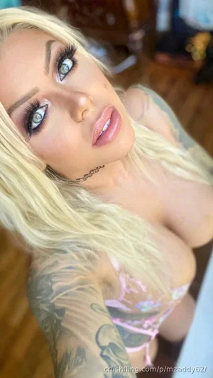 Blonde woman with tattoos in pink lingerie, indoor selfie.