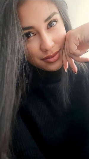 Woman with long dark hair wearing black turtleneck, resting hand on cheek indoors.
