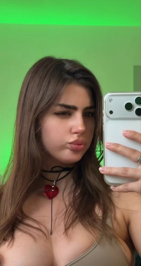 Woman with long brown hair wearing a beige top and red heart choker, taking mirror selfie with