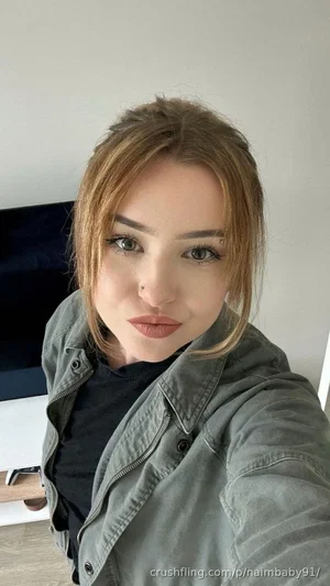 Woman with light brown hair in a green jacket and black top, taking a selfie indoors.