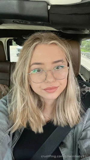 Woman with blonde hair and glasses in a car, wearing a black top and gray jacket.
