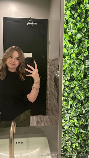 Woman with shoulder-length hair in black top and green pants takes mirror selfie in bathroom with