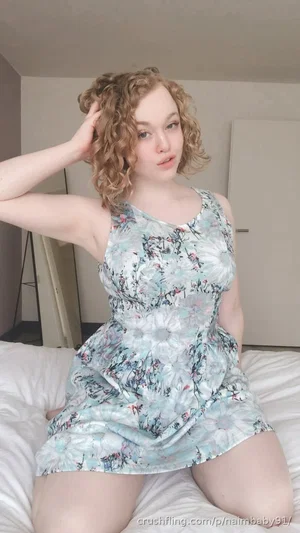 Woman with curly hair in floral dress sitting on bed indoors.