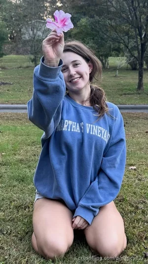 Woman in blue sweatshirt kneeling on grass, holding pink flower, outdoor setting.