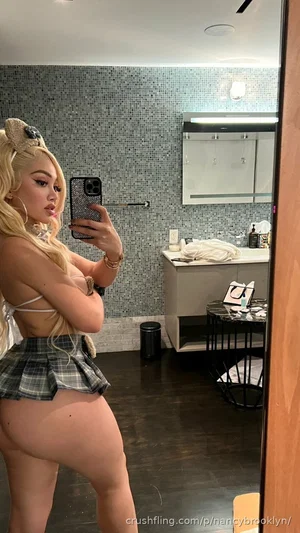 Woman with blonde hair in a plaid skirt and white top takes mirror selfie in bathroom.
