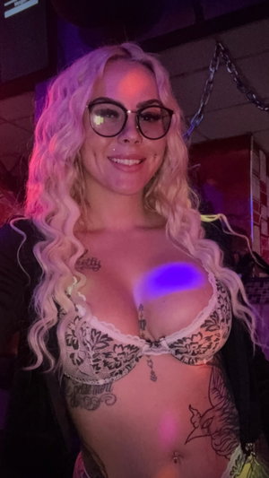 Woman with blonde curly hair, glasses, tattoos, wearing lace bra in dimly lit indoor setting.
