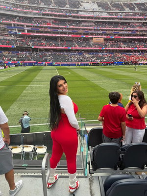 Woman in red and white outfit posing at stadium event, long black hair, smiling.