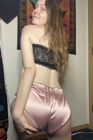 Woman in black lace top and pink satin shorts posing indoors, back to camera.