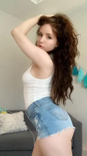 Woman with long brown hair in white tank top and denim shorts posing indoors.