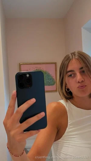 Woman with short hair in white top taking mirror selfie indoors.