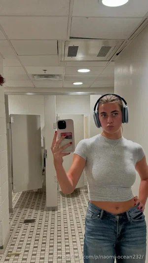 Woman in gray crop top and jeans takes mirror selfie in tiled hallway.