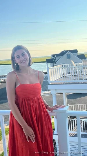 Woman in red spaghetti strap dress standing on deck by water, smiling.