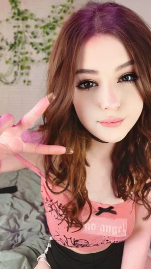 Woman with wavy brown hair in pink crop top, making peace sign indoors.