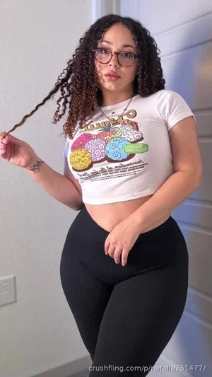 Woman with curly hair wearing a white crop top and black leggings indoors.