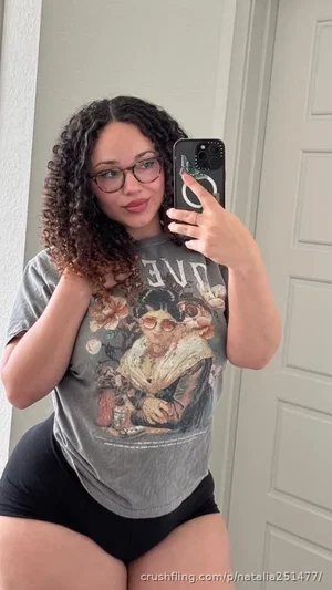 Woman with curly hair and glasses wears graphic tee and black shorts, taking mirror selfie indoors.