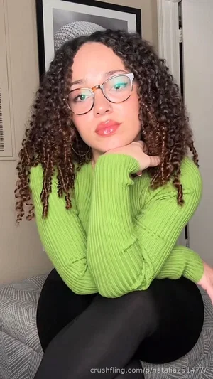 Woman with curly hair wearing green sweater and black tights, sitting indoors.