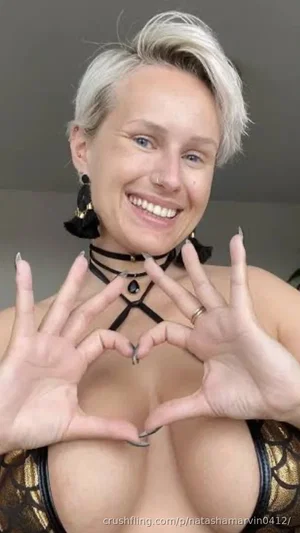 Woman with short blonde hair in a gold and black top, making a heart shape with her hands indoors.