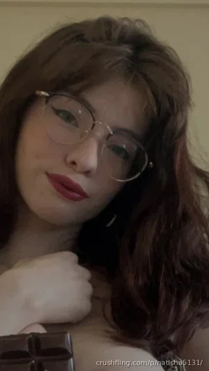 Woman with wavy brown hair and glasses, wearing dark top, holding hand near chest in indoor setting.