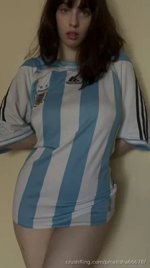 Woman in blue and white striped soccer jersey, standing indoors against plain wall.