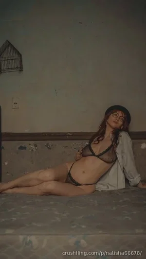 Woman with long hair and glasses in black lingerie and open shirt, lying on bed indoors.