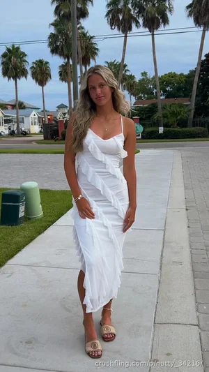 Woman in white ruffled dress standing on sidewalk with palm trees in background.