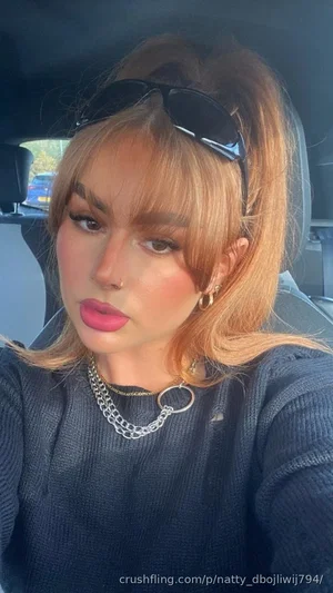 Woman with blonde hair and pink lipstick wearing a dark sweater in a car.