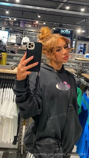 Woman in black hoodie and fanny pack taking mirror selfie in clothing store.