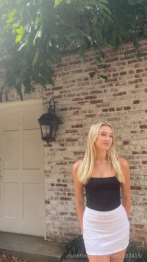 Blonde woman in black top and white skirt standing by brick wall outdoors.