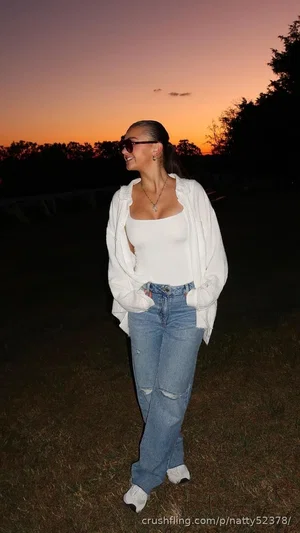Woman in white top and jeans standing outdoors at sunset, smiling.