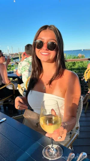 Woman with long dark hair in white strapless top holding wine glass outdoors by water.