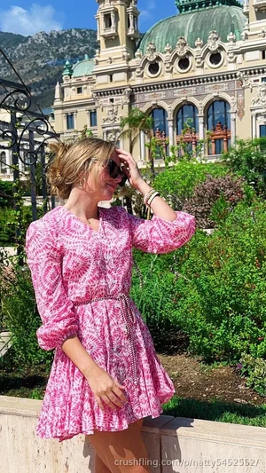 Woman in pink patterned dress standing outdoors near ornate building and greenery.