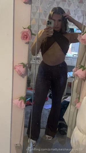 Woman in black crop top and shiny pants poses in mirror selfie in bedroom.