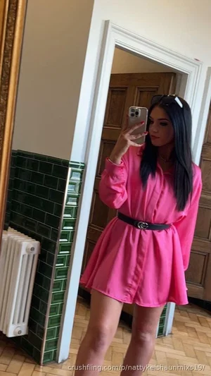 Woman in pink shirt dress with black belt, taking mirror selfie indoors.