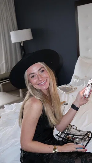 Woman with blonde hair and black hat sits on bed in black dress, holding phone.
