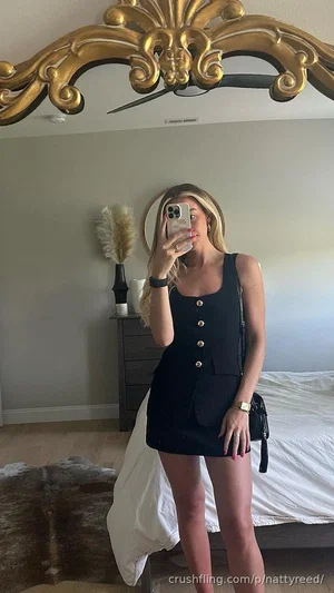 Woman in black sleeveless dress taking mirror selfie in bedroom.