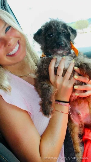 Blonde woman in pink top holding small dog in car.
