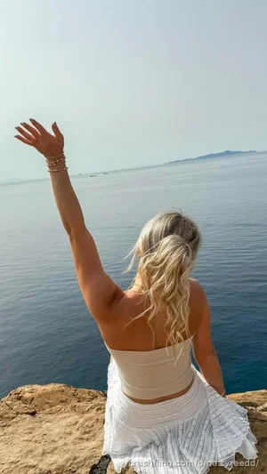 Woman with blonde hair in white top and skirt sitting on rocks by the sea, arm raised.