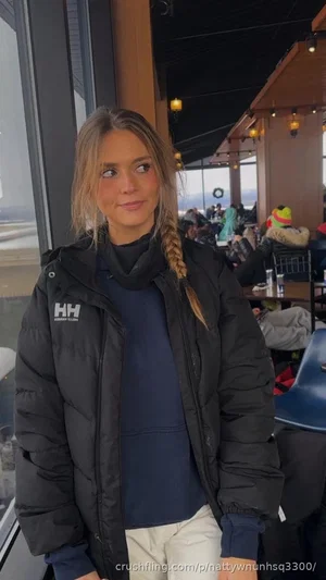 Woman in black puffer jacket and blue top, standing indoors near window in casual setting.