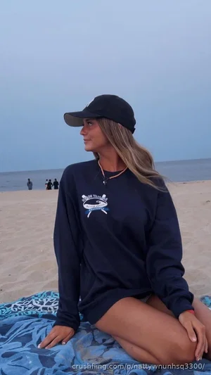 Woman in navy long-sleeve shirt and cap sitting on beach towel, facing side.