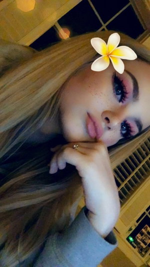 Blonde woman with flower in hair posing for selfie indoors.