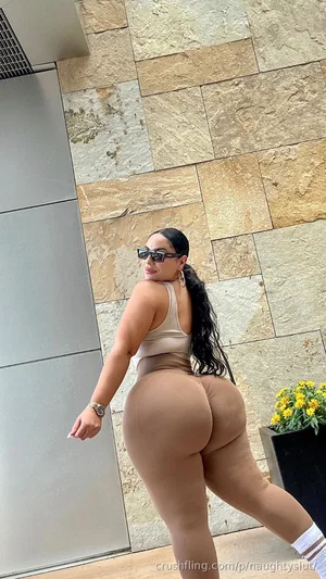 Woman in beige tank top and matching pants posing outdoors near stone wall.