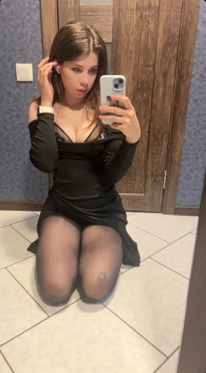 Woman in black dress and tights taking mirror selfie indoors.