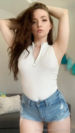 Woman with long brown hair in white top and denim shorts, posing indoors.