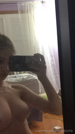 Woman with bare chest and necklace taking mirror selfie in bedroom.