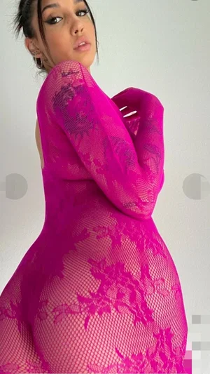 Woman in pink lace bodysuit posing indoors, curvy figure, dark hair tied back.