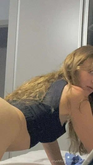 Woman with long blonde hair in dark top, leaning forward on bed indoors.